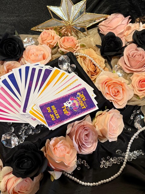 Le jeu De la mort qui tue, cartes et règles Cartes du jeu De la mort qui tue déployées en éventail, avec le livret de règles, mis en scène avec des roses